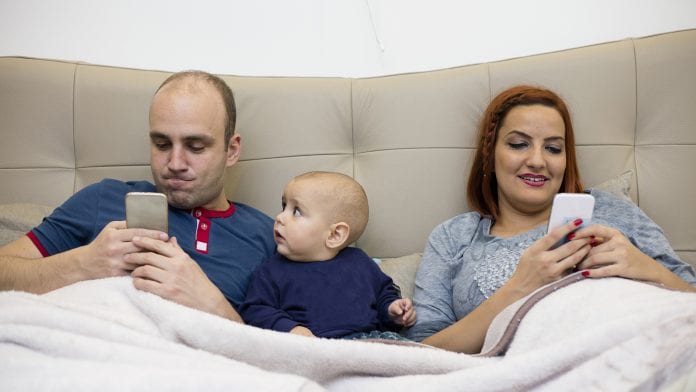 Parents using phones white lying in bed with baby