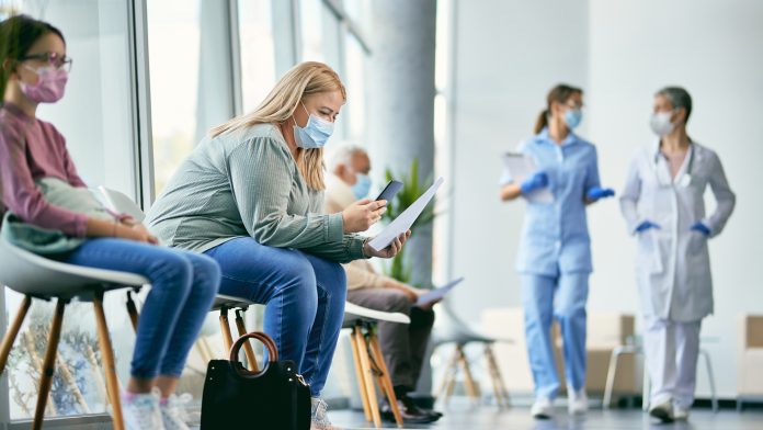 Woman with face mask using smart phone while reading her medical report in waiting room at the clinic. NHS waiting lists