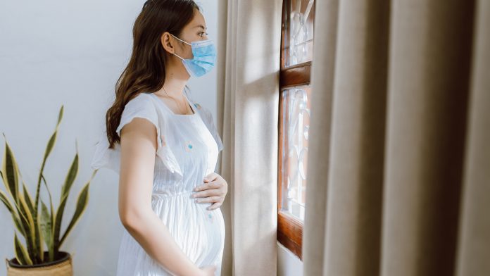 Young pregnant Asian woman wearing medical mask standing near window at home for self quarantine from spread of Covid 19 diseases. asymptomatic Covid