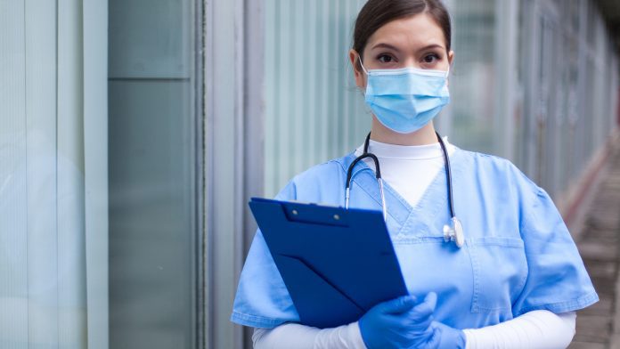 Female GP UK doctor holding blue clipboard standing outside hospital or clinic NHS staff